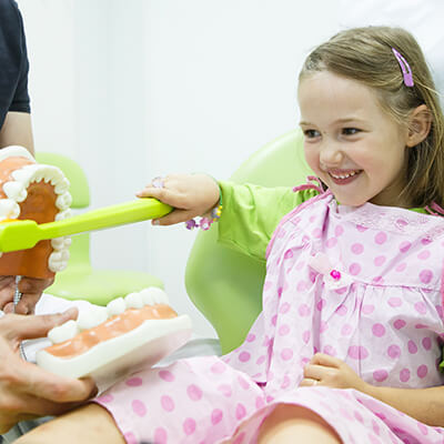 A girl wearing a pink dress inside the dental office brushing some false teeth with a green toothbrush