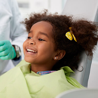 A girl getting services in the dentist's chair while smiling and wearing a green bib